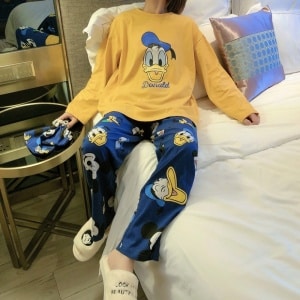 Söt Disney Donald Pyjamas för kvinnor i senapsgult och blått som bärs av en kvinna som sitter på en säng i ett hus