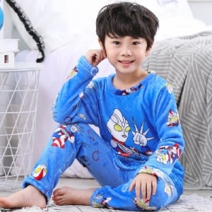Blå pojks superhjälte pyjamas i flanell som bärs av en liten pojke som sitter på en matta framför en säng i ett hus