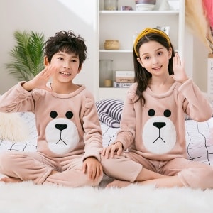 Bruna fleecepyjamas för barn som bärs av en liten flicka och en liten pojke som sitter på en matta i ett hus